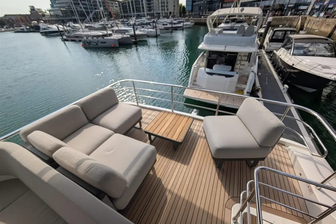 Modern boat deck featuring grey seating, a wooden table, and moored yachts reflecting in calm water.